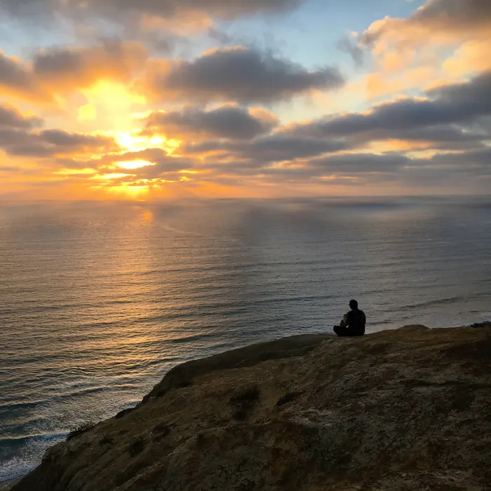A person sitting quietly on a cliff above the ocean at sunset